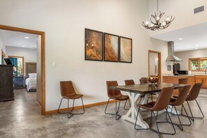 A modern dining area with a large table and chairs, a chandelier, and artwork on the wall. - Roadrunner | Riverfront 3BR with Views & Fire Pit (Leakey)