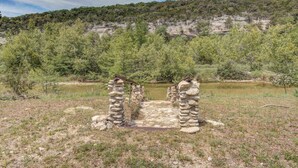 Estructura de piedra con una viga de madera, rodeada de árboles y una colina al fondo.