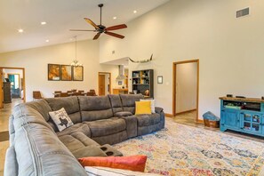 A spacious living room with a sectional sofa, a ceiling fan, a decorative wall piece, and a bookshelf.