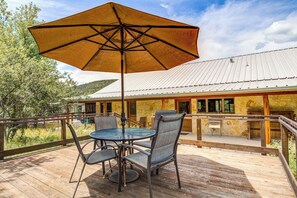 A wooden deck with a table and chairs, an umbrella, and a building with stone walls and a metal roof in the background. - Roadrunner | Riverfront 3BR with Views & Fire Pit (Leakey)