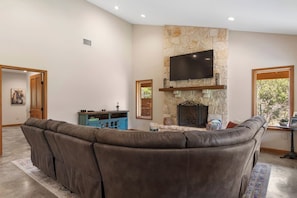 A living room with a stone fireplace, a flat-screen TV, and a large L-shaped sectional sofa. - Roadrunner | Riverfront 3BR with Views & Fire Pit (Leakey)