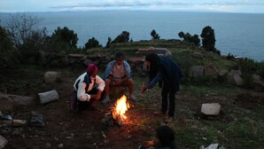 Tres personas alrededor de una fogata en un entorno natural con vista al mar.