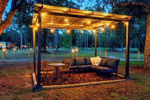 Un espace de détente abrité, avec un canapé et des fauteuils, entouré d’une pergola en bois agrémentée de guirlandes lumineuses, situé dans un endroit boisé.