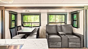 A modern RV interior with a dining area, a sofa, and large windows offering a view of the outdoors. - Fuel Your Passion for Epic Experiences and Outdoor Adventures at the Moto Ranch SX! (Brooksville)
