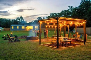 Un espace extérieur aménagé avec une terrasse couverte, des guirlandes lumineuses, un foyer et une plateforme en bois.