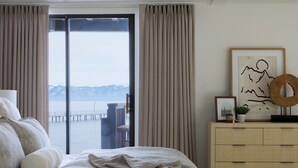 Ein Schlafzimmer mit einem großen Bett, einer Kommode, einem gerahmten Bild und Blick auf ein Gewässer durch eine Schiebetür.