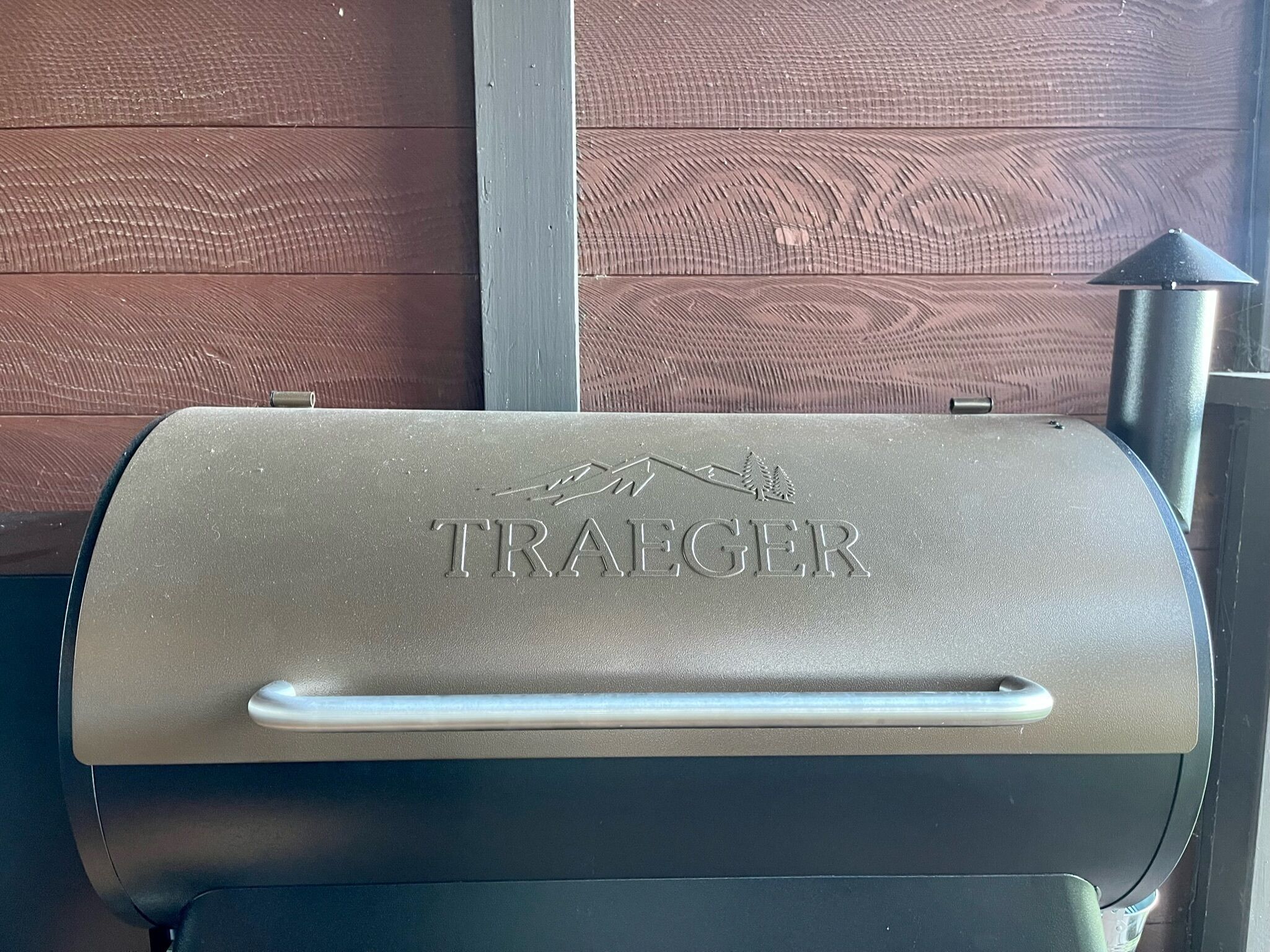 A Traeger grill with a handle and a logo, set against a wooden wall.