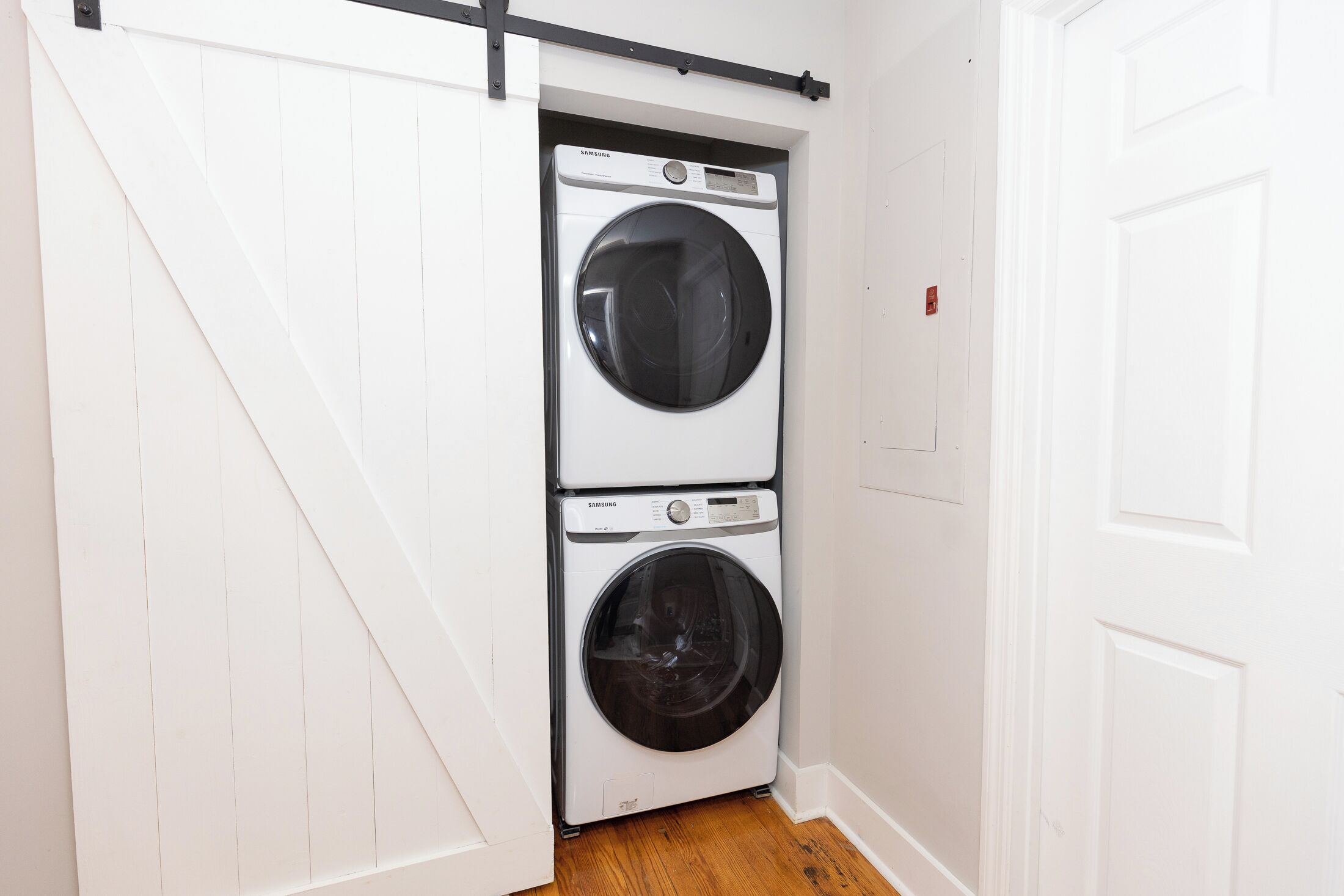 A laundry room with two stacked front-loading washing machines, a wooden floor, and white walls.
