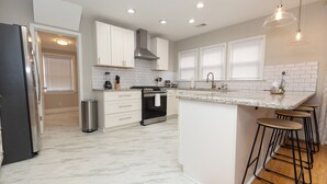 A modern kitchen with white cabinets, a stainless steel refrigerator, a built-in oven, and a bar area with stools. - Bexley Place (North Charleston)
