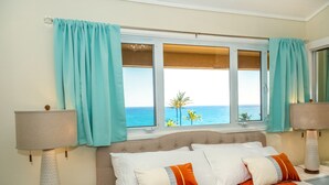 A bedroom with a bed, two bedside lamps, a window with curtains, and a view of the ocean. - Remodeled Family-Size Condo with Fantastic View of the Ocean and Pool (Koloa)