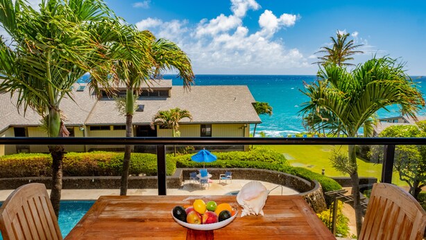 A wooden table with a fruit bowl and a seashell, overlooking a pool and a golf course with a view of the ocean. - Remodeled Family-Size Condo with Fantastic View of the Ocean and Pool (Koloa)