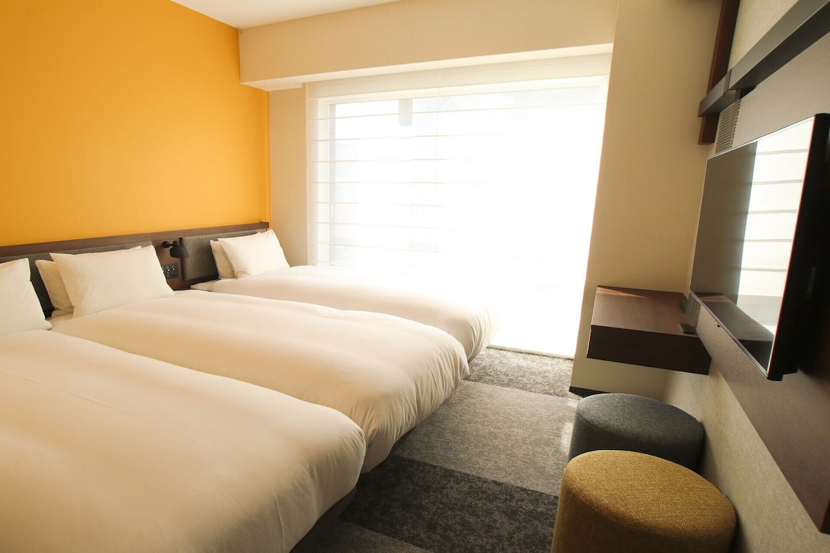 A hotel room with two beds, a TV, a window, and a small seating area.