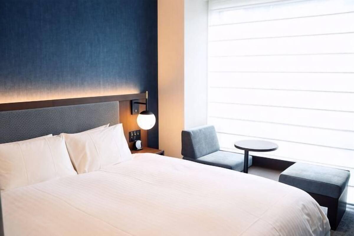 A hotel room with a large bed, a bedside table with a lamp, a small round table, a chair, and a window with blinds.