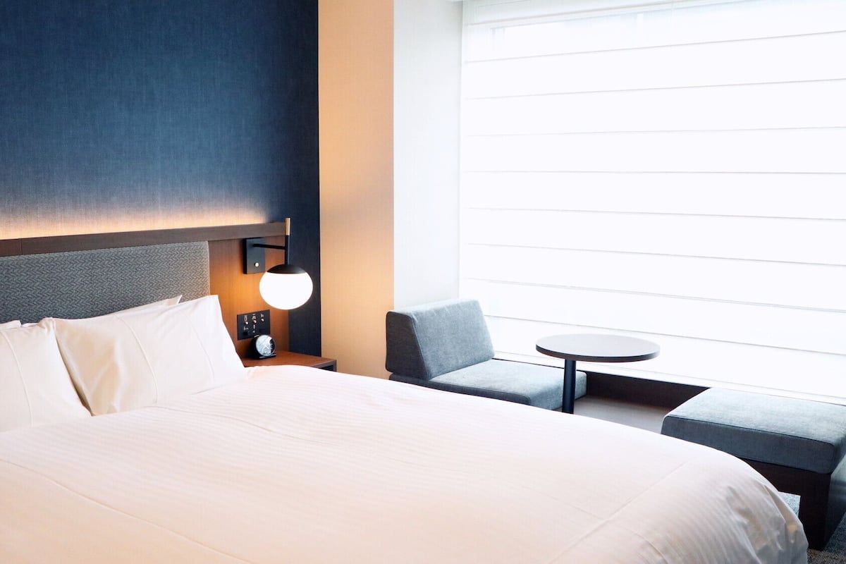 A hotel room with a large bed, a small table, a chair, and a bedside lamp.