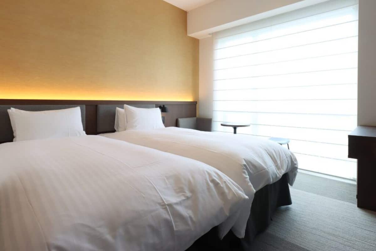 A hotel room with a large bed, a bedside table, a small table, and a window with blinds.