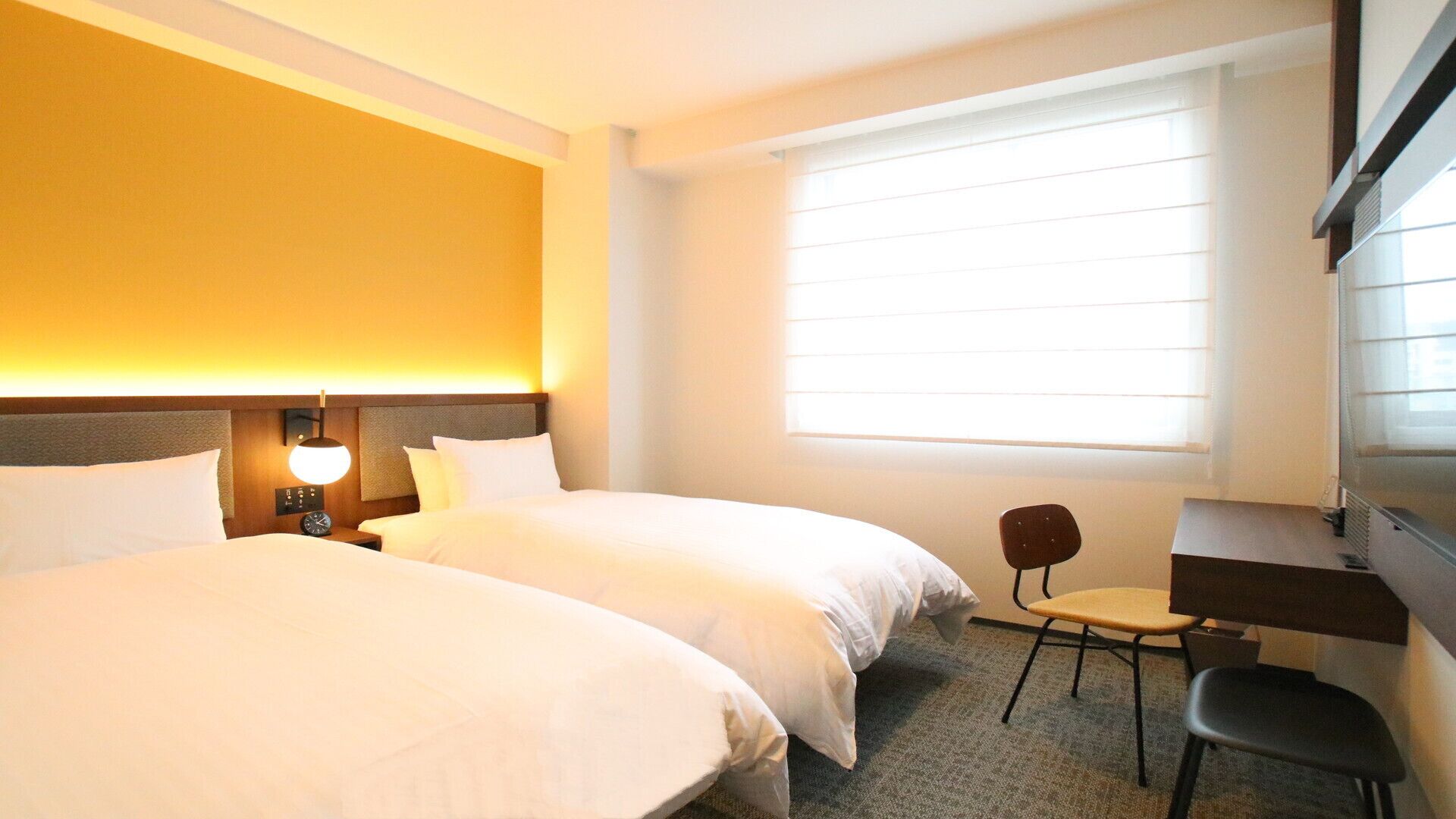 A hotel room with two beds, a desk, a chair, a lamp, and a large window with blinds.