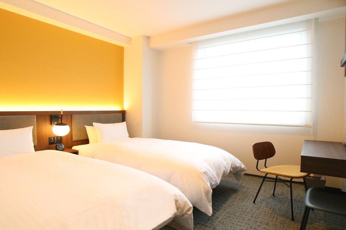 A hotel room with two beds, a desk, a chair, a lamp, and a large window with blinds.