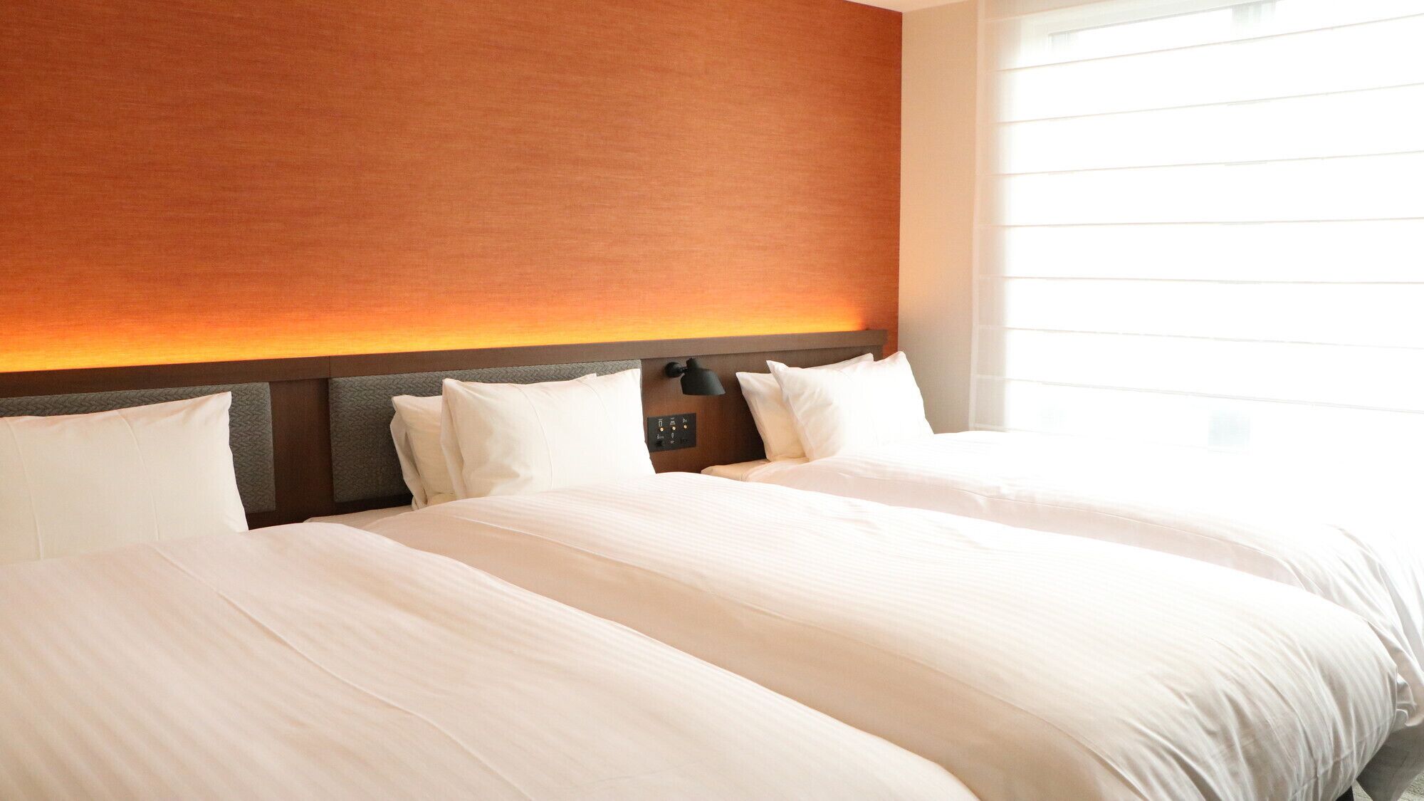 A hotel room with two beds, a headboard with a light fixture, and a window with blinds.