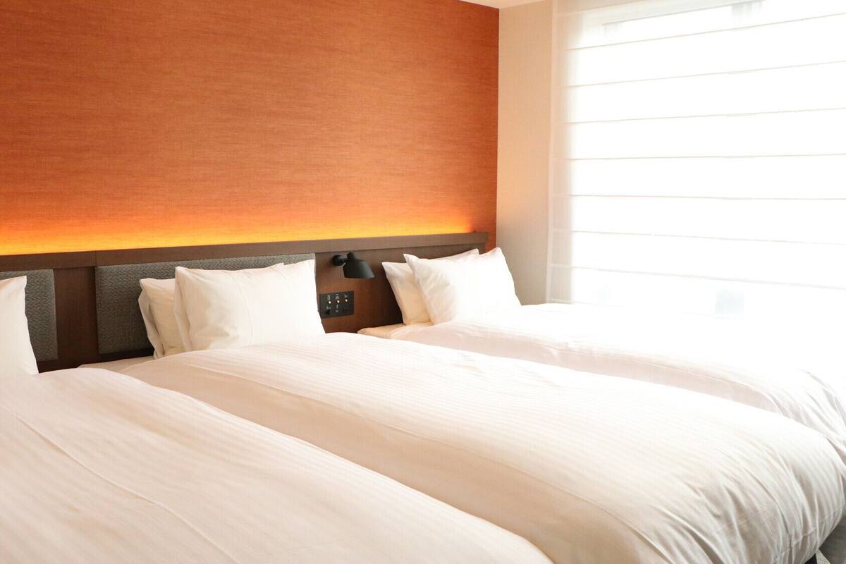 A hotel room with two beds, a headboard with a light fixture, and a window with blinds.