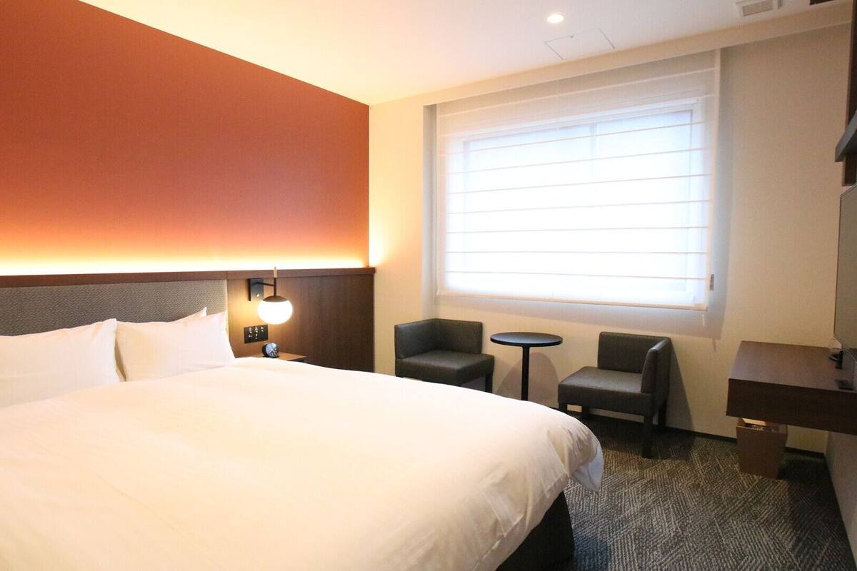 A hotel room with a bed, a desk, a chair, a small table, and a TV.