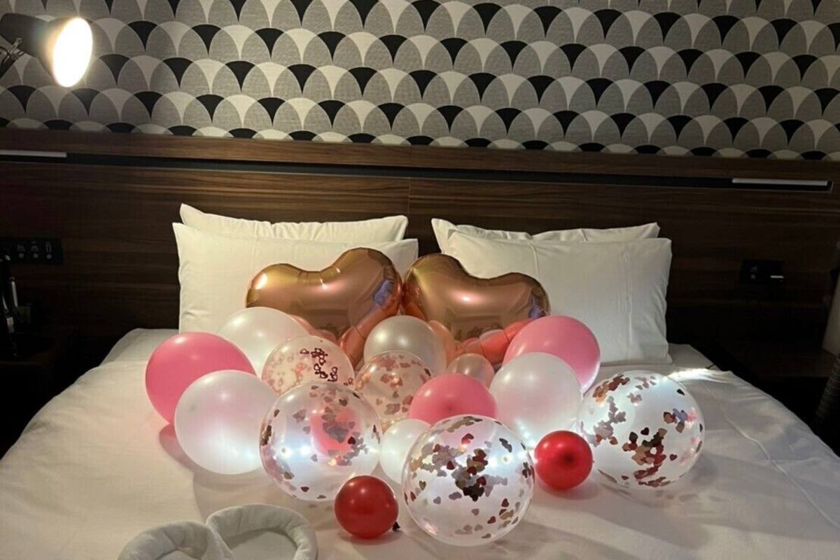 A bed with a heart-shaped pillow, a heart-shaped balloon, and a heart-shaped balloon on a white bedspread.