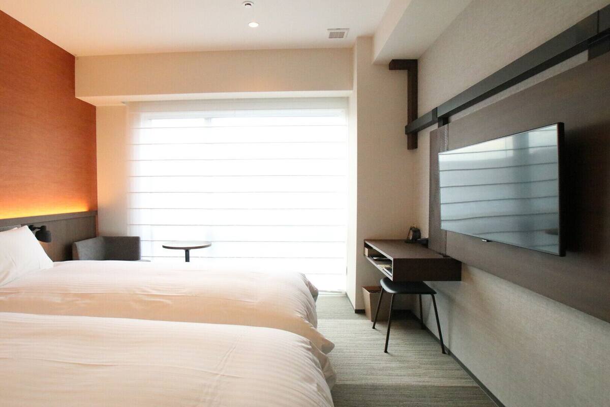 A hotel room with a bed, a desk, a chair, a TV, and a window with blinds.