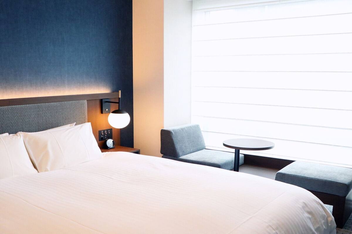 A hotel room with a large bed, a small table, a chair, and a window with blinds.
