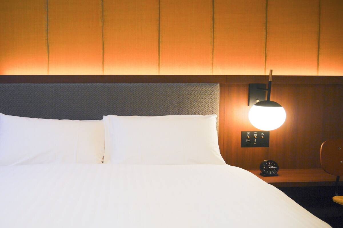 A hotel room with a bed, a nightstand with a clock, and a wall-mounted lamp.