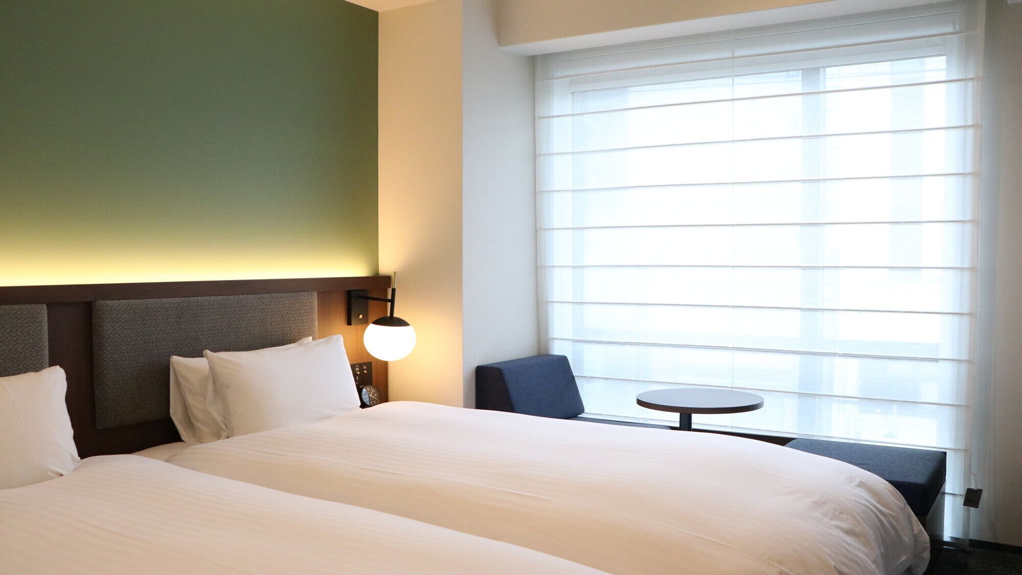 A hotel room with two beds, a small table, a chair, and a large window with blinds.