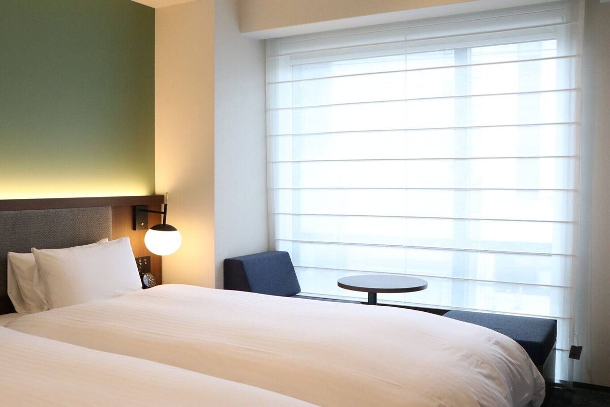A hotel room with two beds, a small table, a chair, and a large window with blinds.