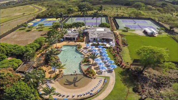 An aerial view of a resort with a central swimming pool, tennis courts, and a landscaped area with umbrellas.
