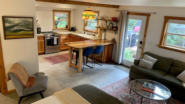 A cozy living room with a sofa, a coffee table, and a rug. There's a kitchen area with wooden cabinets and a dining table with chairs.