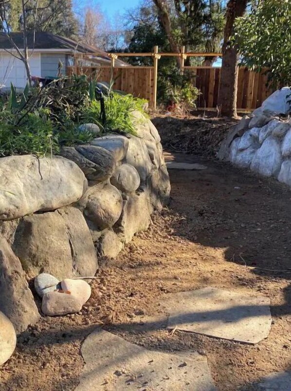 Un patio trasero con un muro de piedra, cerca de madera y un área de jardín con plantas.