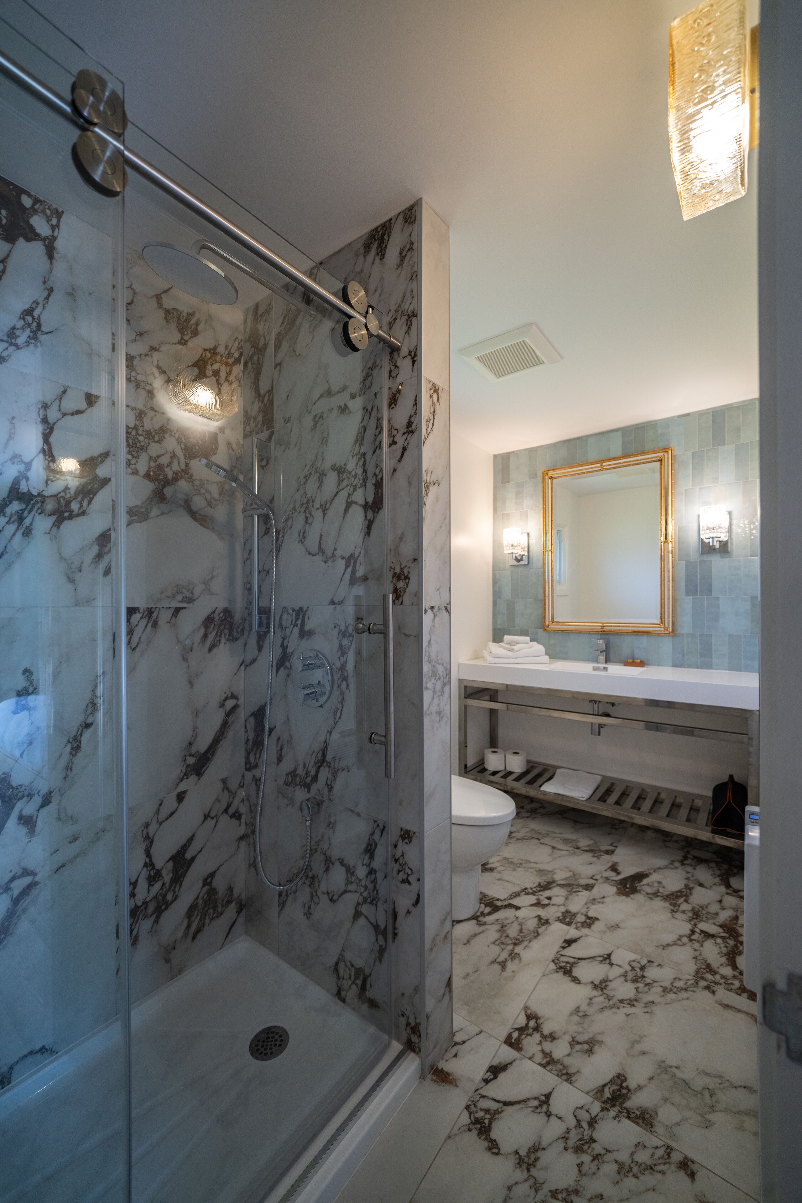 A bathroom with a glass-enclosed shower, a marble-tiled floor, a white sink, and a mirror with a gold frame.