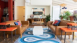 A spacious living area with a brown leather sofa, a white coffee table, and a blue patterned rug. There are two orange side tables and a bookshelf with red vertical panels. - Motel Chelsea (Chelsea)