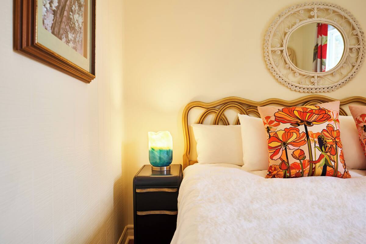 Ein ordentlich bezogenes Bett mit floralen Kissen, ein Nachttisch mit Lampe und ein runder Spiegel an der Wand.