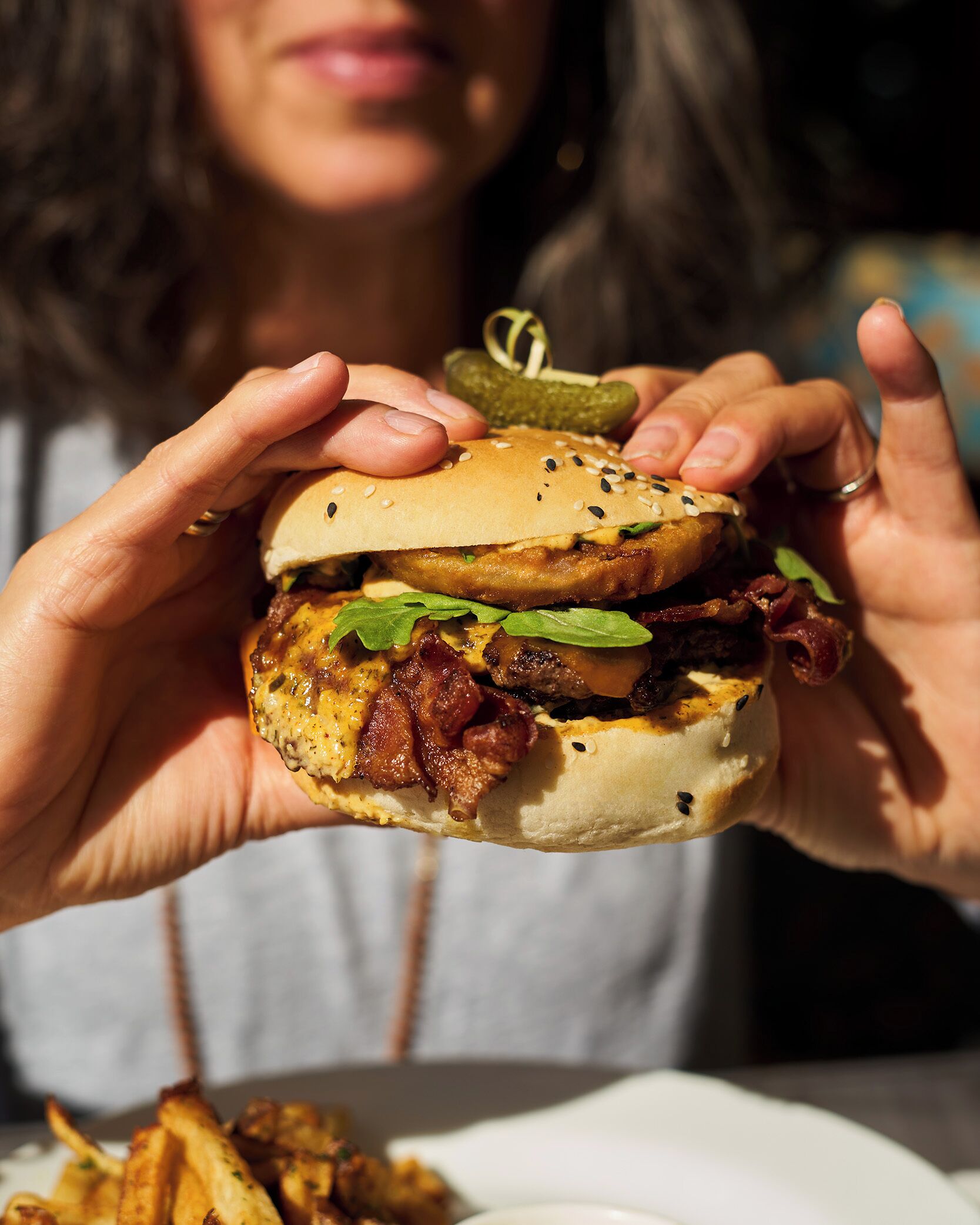 Eine Person hält einen Burger mit sichtbaren Zutaten wie Salat, Speck und einem Sesambrötchen.