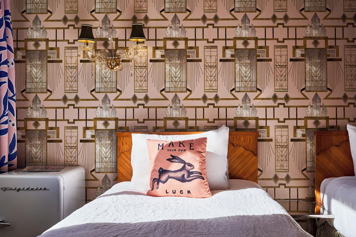 Ein Hotelzimmer mit gemustertem Wandpapier, einem weißen Bett mit einem Kissen mit einem Kaninchenmotiv und einem Kühlgerät im Vintage-Stil.