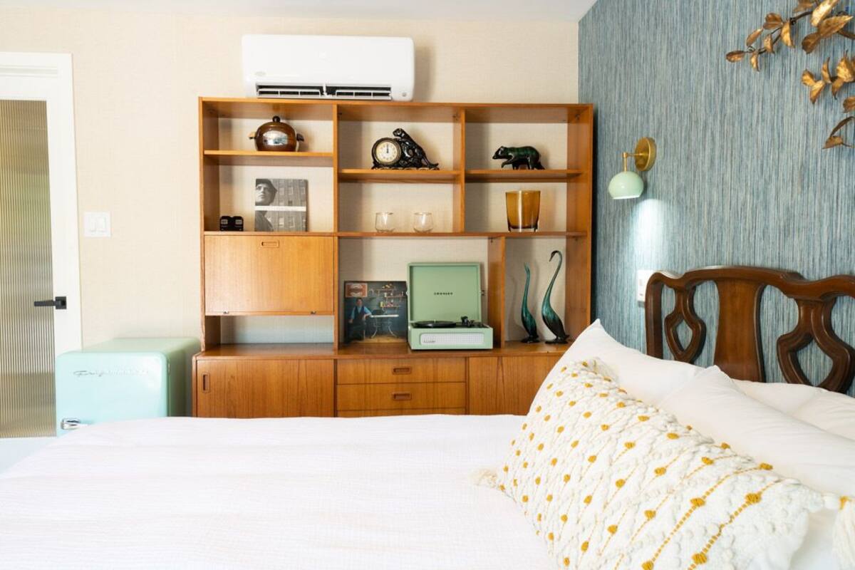 Ein Schlafzimmer mit einem Bett, einer Holzkommode mit diversen Gegenständen, einem Vintage-Radio und einer Wand-Klimaanlage.