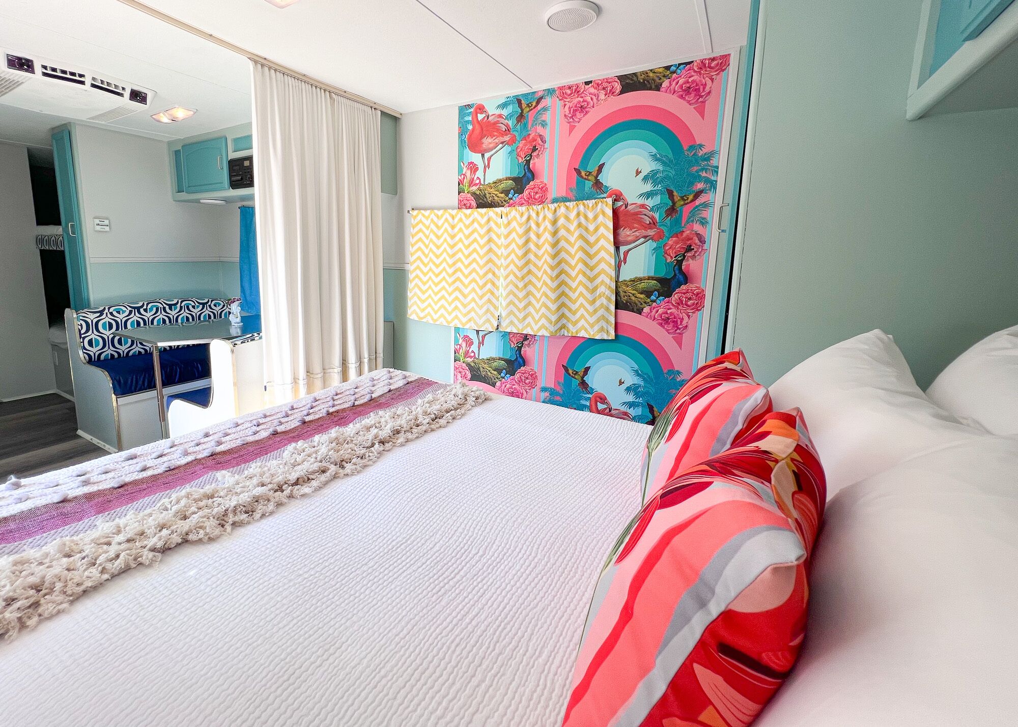 A bedroom with a bed, a window with curtains, a wall with a colorful mural, and a small table with a chair.