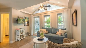 Ein geräumiges Wohnzimmer mit hohen Decken, einem Deckenventilator, einem großen Fenster mit Vorhängen, einem grauen Sofa mit dekorativen Kissen, einem weißen Couchtisch, einem Korbstuhl und einem Bücherregal mit gerahmten Bildern.