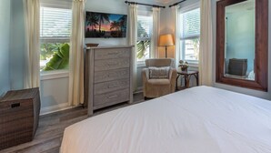 A bedroom with a large bed, a dresser, a chair, a lamp, and a painting of a sunset. - Campbell Suite (Key West)