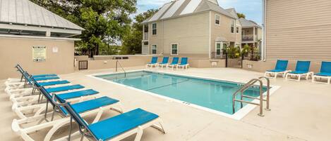 A pool area with lounge chairs, a pool, and a building with a sloped roof.