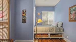 Um quarto com uma cama, um abajur, uma obra de arte na parede e uma janela com persianas.