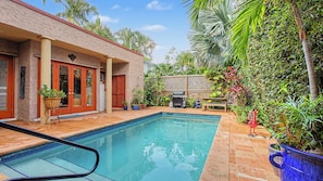 Piscina com deck revestido, área de varanda coberta e diversos vasos com plantas.