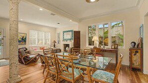 Uma sala de estar ampla com uma mesa de jantar de vidro cercada por cadeiras de vime, uma lareira e uma grande janela com persianas.