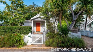 A white house with a red door, surrounded by lush greenery and a white picket fence. - Poinciana House (Key West)