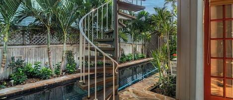A spiral staircase leading to a pool area with a wooden deck and tropical plants.
