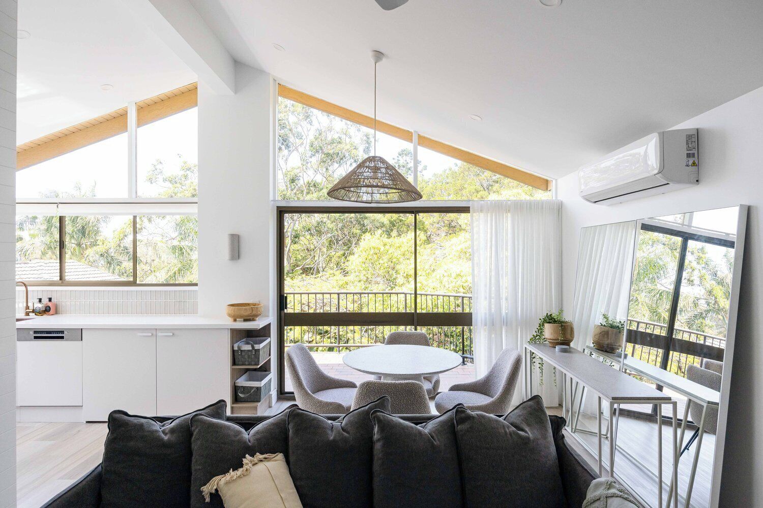 A modern kitchen with white cabinets, a dining area with a round table and grey chairs, and a balcony with a view of greenery.