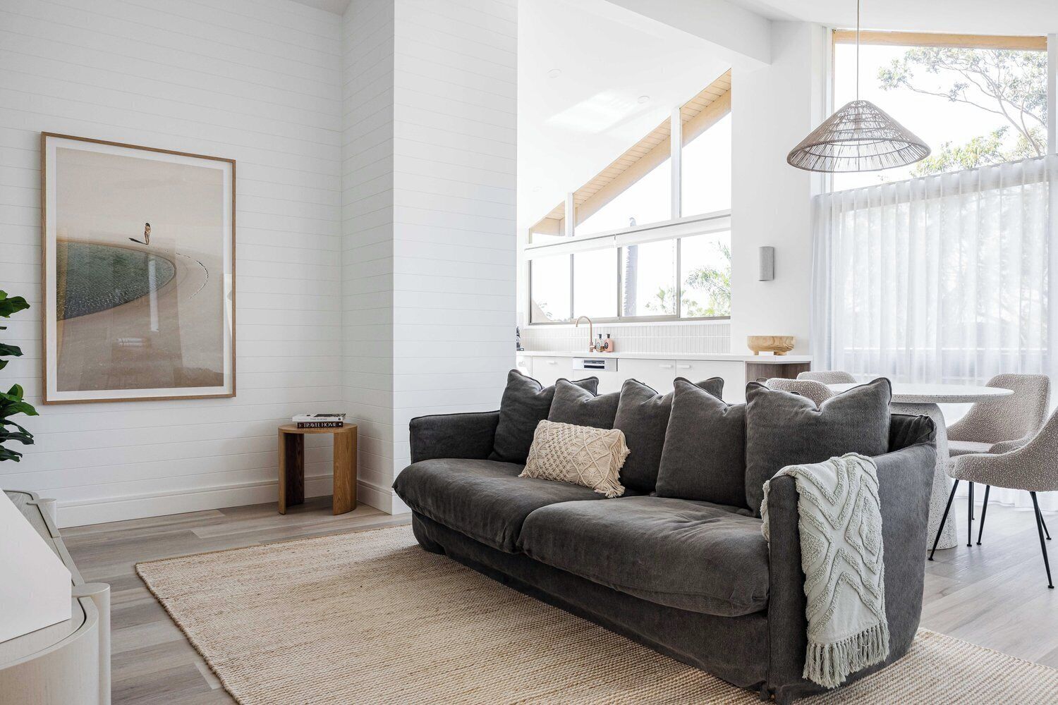 A modern living room with a grey sofa, a small wooden side table, and a large window with sheer curtains.